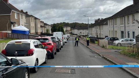 A residential street with beige buildings with dark rooves. They have fenced off front gardens and cars are parked along the street. A police cordon blocks off the street and police officers can be seen further down the road