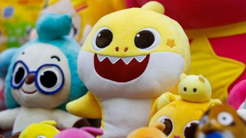A row of Pinkfong's Baby Shark character soft toys on a display. The main yellow Baby Shark sits at the centre.