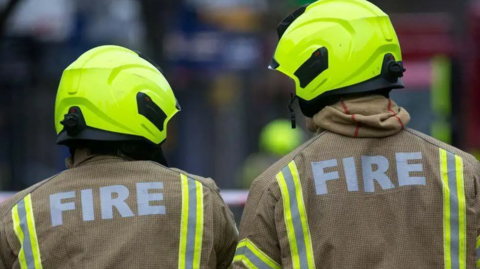 Two firefighters in uniform stand with their backs to the camera. Both have helmets on with the word "fire" written on their back.