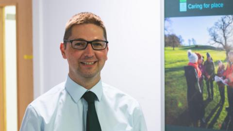 Tewkesbury Borough Council leader Richard Stanley is standing in an office. He is wearing a light blue shirt and a black tie. He is smiling at the camera.