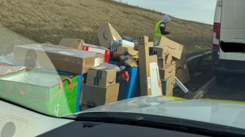 A big pile of boxes by the side of a road, with a man, wearing a high-visibility jacket, with his hood up, in the distance. The parcels are by a grass verge with a white van to the right. 