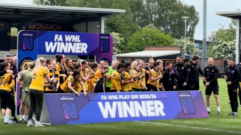 The players and team staff gather behind a banner on the pitch which says "FA WNL play off 2025/26 winner" 