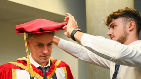 Bearded man wearing a white shirt and blue tie fits a red hat on to Kieran McKenna. The Ipwich manager is wearing a red and yellow academic robe, a white shirt, with the top button unfastened, and dark tie