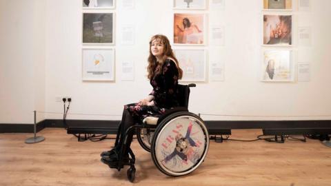 Wheelchair user Hope Caitlin. The chair's wheels have a Taylor Swift-themed image. Behind her on a white wall are the artworks she has created for the exhibition.