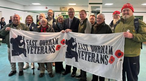 Group of men and women standing holding a banner in a large room which reads "East Durham Veterans".