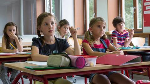 Pupils sit in a classroom and listen attentively. They have files and paper on the desk and pencil cases. The classroom has large windows and there is a notice board.