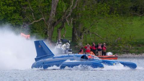 Bluebird in action on Coniston Water. It is a bright blue rocket-shaped craft. Spray is shooting from the rear where the jet engine is blowing the surface water. Meanwhile water is bubbling up around the craft's front and sides. There are support boats closer to the shore and four people are watching through binoculars. 