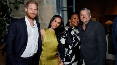 Harry and Meghan smiling next to two others, wearing cocktail attire, at a house