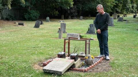 A man stands with head bowed at a grave. The headstone is laid flat as if it has been knocked over. It is surrounded by short grass and other, standing headstones. The man wears a grey jumper, jeans and glasses.