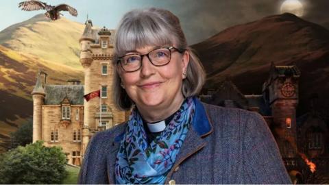 A woman with grey bobbed hair and glasses smiles at the camera. She is wearing a blue patterned blouse, blue blazer and a dog collar. In the background are hills and a castle, with an owl flying in the sky. 