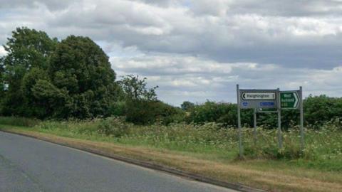 A view of the hedgerow and sign on the A68 with a sign pointing to Heighington in one mile.