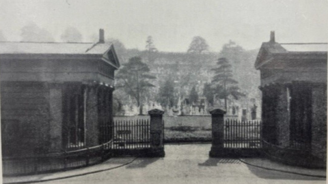 An historic image of the gates in black and white. The gates are central with the gatehouses either side and the cemetery behind, with trees and grass there.
At the front of the picture, it is paved.