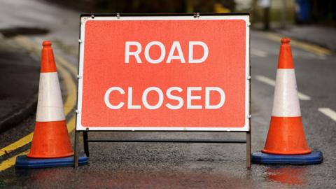 A red road closed warning sign has been placed on a road. Traffic cones are either side of the sign. 