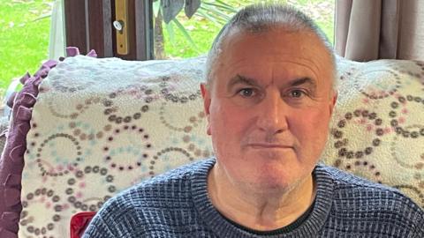 A man in his 60s sits on a sofa in a living room. He has short grey hair and is wearing a blue woollen jumper. The sofa is covered in a white throw with a pink, blue, purple and grey pattern.