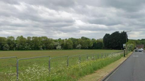 A field next to a road. The field has a metal fence blocking it off. Trees are seen in the distance. 