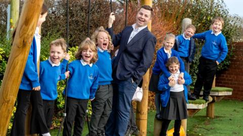 Ben McAuley and a number of primary-age pupils stand on wooden children's play equipment, in a playground. Mr McAuley wears a navy blue suit and a white shirt. He has short brown hair. The pupils wear blue sweatshirts, black trousers or skirts and white polo shirts.