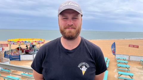 A blond man with a beard, and wearing a light purple baseball cap. He is wearing a dark navy shirt and standing by a beach.