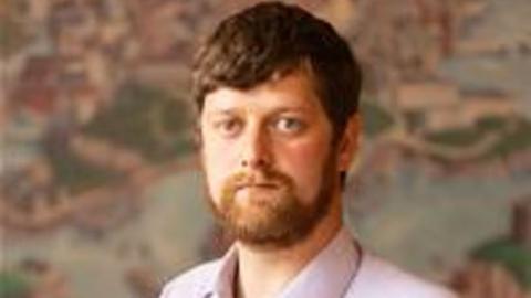 Ben Rowe in a portrait photograph taken by Plymouth City Council. He has brown hair and a beard, and is wearing a pink shirt.