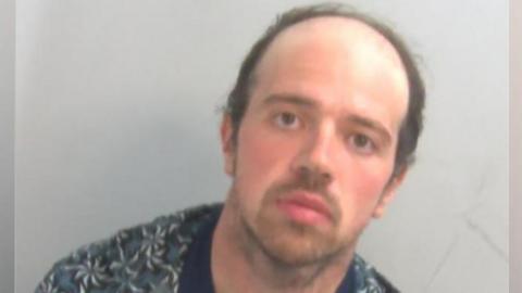 A police custody mugshot of Luke D'Wit, who is wearing an unbuttoned shirt with a T-shirt underneath. He has short dark hair and a blank expression on his face.