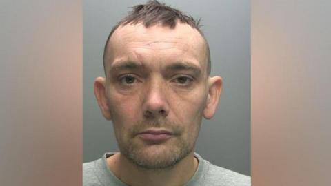 A mugshot of Craig Weaver. He has short brown hair and a receding hairline. He has brown to green eyes and stubble. He is wearing a grey sweater and is looking at the camera with a serious expression