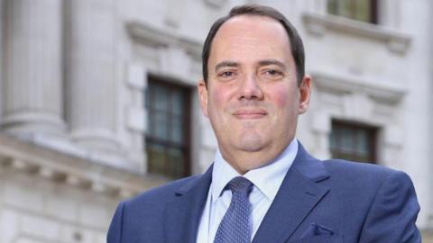 Marco Salzedo smiles for the camera. He is wearing wearing a blue suit and tie with a white shirt. He is clean shaven with short brown hair and is standing in front of a white stone building with wooden window frames.
