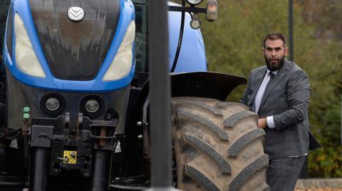 Luke Poots is wearing a grey suit and is standing next to a blue tractor. He has a dark brown beard and brown hair.