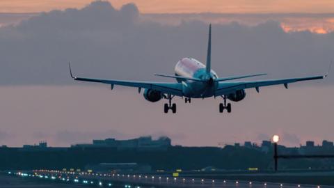 A plane taking off above a runway with the sun setting in the cloudy sky above it.