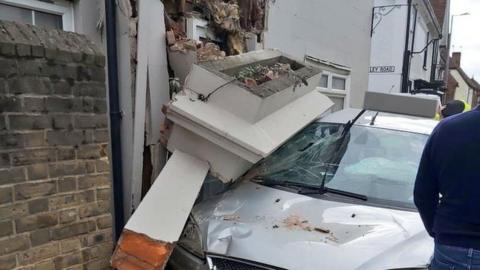 A silver car crashed into a white building. Parts of the wall have crumbled on top of the car and exposed brickwork can be seen.