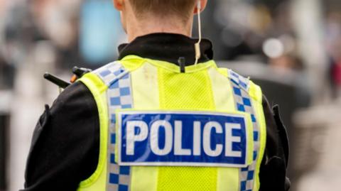 A picture of the back of a police officer. He is wearing a black police uniform with a bright yellow vest over the top. On the vest is a blue strip with the words police written in white letters.