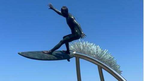 A bronze statue of a surfer wave riding. Its dark and taken on a bright sunny day with blue skies in Newquay.