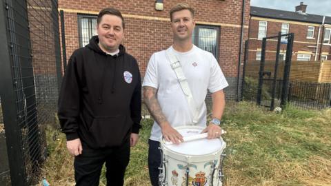 Two men look at the camera smiling. The man on the left is dressed in black trousers with a blacl hoodie and has black hair. The man on the right is wearing a white tshirt with blonde hair, his lower half is covered by a white drum with white drumsticks on top. 