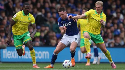 A footballer wearing a blue and white kit looks down at a football as a player to his left, wearing yellow and green, goes in for a tackle. A second player in yellow and green approaches from the right. Spectators watch from the stands in the blurred background. 