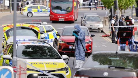 A forensic officer stands in a pale blue protective suit surrounded by police vehicles. A group of police officers can be seen in the background