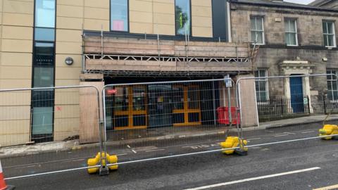 Durham Bus Station in Durham. The entrance has wooden scaffolding covering the building and looks like it is awaiting repairs. There is large metal fencing protecting the entrance.