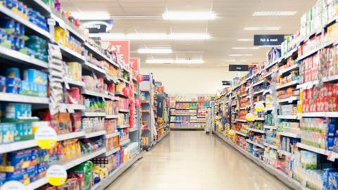 Shelves in a grocery store aisle.
The products are blurred out but colourful and the titles at the top of the aisle are blue and "fish and meat" sign is visible. 
The floor is cream and the lights are bright. 