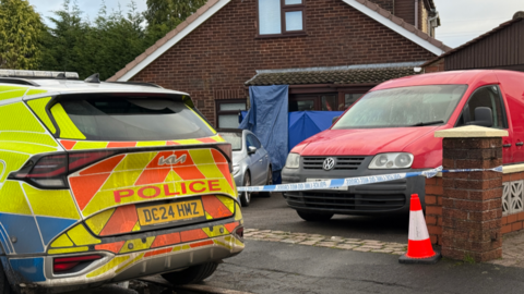A police car is parked outside the drive of a dormer bungalow. There is a van and car parked on the drive, with police tape across the entrance. Blue sheeting hangs above the door of the property