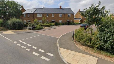 Road leading into residential street with terrace of new homes along tree lined paths - it is a dry and sunny day.