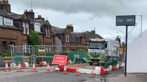 A road closure outside a chip shop - an area of the road has been fenced off with work taking place on the ground. There is a lorry within the work area.