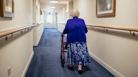 An elderly woman walks down a corridor in a care home with her back to the camera. She is using a walker for support and is wearing a blue cardigan and a long blue flowery skirt. The walls are cream and the carpet is blue.