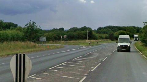 A road with a junction turning off to the left. In the foreground is the back of a small road sign. On the right-hand carriageway, a white van and an HGV can be seen. Trees and hedges line both sides of the road.