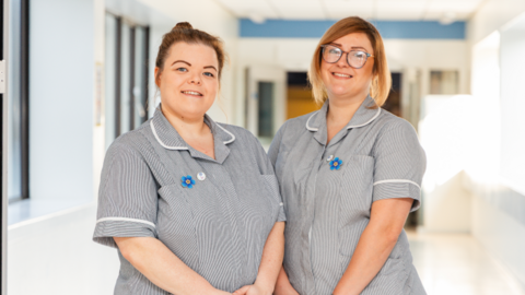 Two women in grey nurses' scrubs stand together side by side and smile at the camera. They both have a blue dementia pin in the shape of a flower on their scrubs.