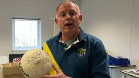 Simon Clarkson, who has short grey/brown hair and is wearing a blue and yellow Carlisle United top, is holding up an old football which is signed in pen.