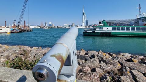 A sunny scene of Gosport looking out over the harbour to Portsmouth. A old-style cannon is seen in the foreground with the Gosport ferry in the harbour. The Spinnaker Tower can be seen in the distance under a clear blue sky.