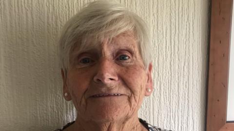 An elderly woman with white short side fringe hair and pearl earrings on against a white wall. 