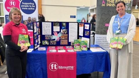 Two women standing holding colourful booklets at either side of a table, covered with a blue tablecloth, with leaflets and posters on it. Heather Gibson is wearing black trousers, a black long-sleeved shirt and a pink shirt. She has shoulder-length blonde hair. Alex Wray is wearing neutral-coloured trousers, a white cardigan and a light blue shirt. She had brown hair tied back. There are posters and logos for Fitmums and Friends.