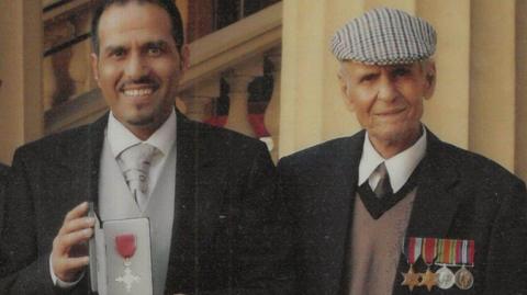 Mashuq Hussain holding his OBE in grey and black tie and tails, standing next to Ghulam Hussain wearing his medals