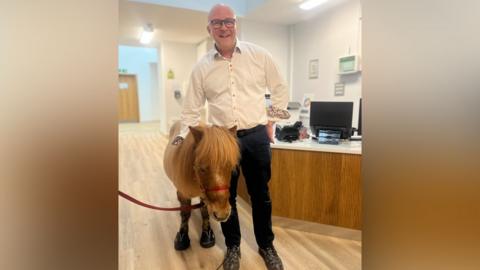 Derek is bald and wearing a white shirt with colourful buttons, and black trousers. Next to him is a small red brown Shetland Pony with a red harness on it. They are in a building's reception area with a computer on a desk behind them.