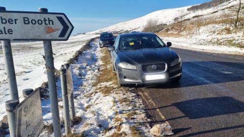 A black car, which has a parking ticket on the windscreen, parked on a double-yellow line near road signs in the Peak District in Derbyshire on a snowy and bright day.