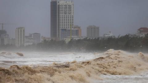 Large swells of waves are seen on the shore in Vietnam and buildings are seen in the background.