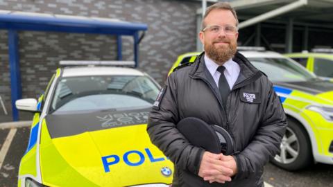 Patrol Insp Wes Watkins standing in front of two police cars. He is a beard and is wearing glasses, a tie and is holding his hands together.